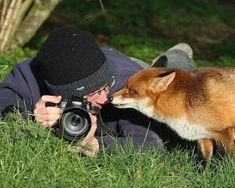 a man laying in the grass with a camera next to a small dog that is looking at him