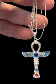 an egyptian cross pendant with blue and red stones on it's side, being held by a hand