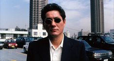 a man wearing sunglasses standing in the middle of a busy street with tall buildings behind him