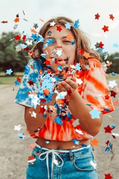 a girl in an orange shirt and blue jeans is blowing out red, white and blue confetti