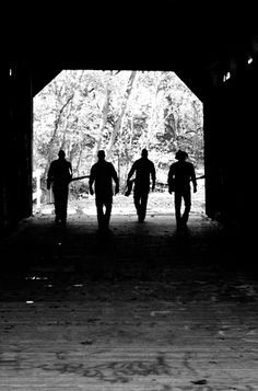 black and white photograph of three men walking out of a covered bridge with trees in the background