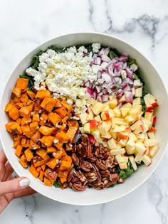 a bowl filled with chopped vegetables, nuts and feta cheese next to someone's hand