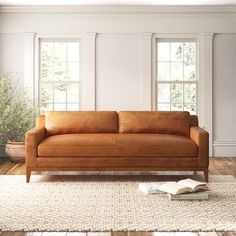 a brown leather couch sitting on top of a wooden floor next to two large windows