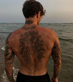 a man with tattoos on his back standing in the water at the beach looking out to sea