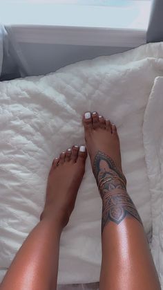 a woman's legs with tattoos laying on top of a white comforter next to a window