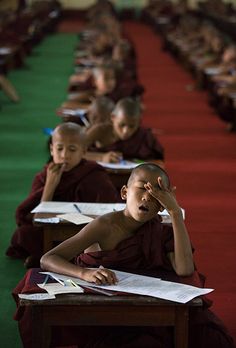 young monks sitting at desks with papers in front of them and one person holding his head