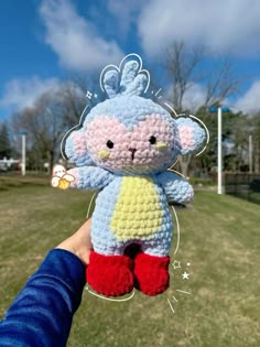 a person holding up a crocheted stuffed animal in the air with grass and trees behind it