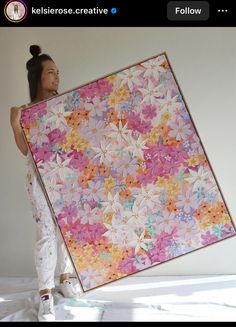 a woman holding up a large quilt with pink and orange flowers on it, in front of a white wall