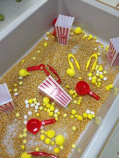 popcorn and candy in a plastic container filled with corn kernels, red spoons, and yellow candies