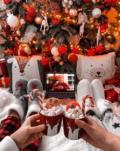 two people are holding cups with marshmallows in front of a christmas tree