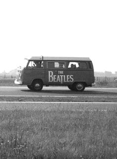 an old vw bus with the beatles written on it driving down a road in front of a grassy field