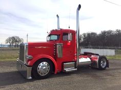 a red semi truck parked in a parking lot next to a fence and grass field