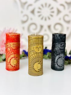 three different colored candles sitting next to each other on a white table with flowers in the background