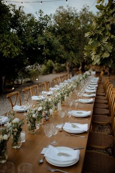 a long wooden table with white plates and place settings on it, surrounded by greenery