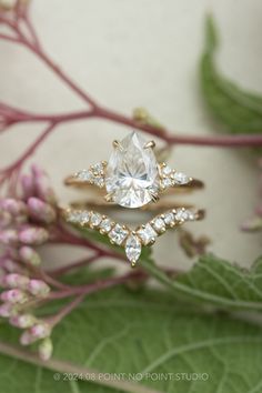 two gold rings with diamonds on them sitting next to some pink flowers and green leaves