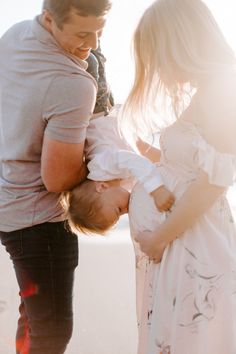 a man holding a baby while standing next to two other people on the beach at sunset