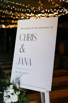 a welcome sign with flowers and lights in the background at a rustic wedding reception venue