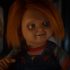 a creepy doll with red hair and blue eyes looks at the camera while standing in front of two other dolls