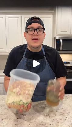 a man in an apron holding a plastic cup filled with food on top of a kitchen counter