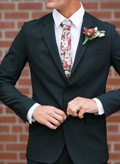 a man in a suit and flowered tie smiles at the camera while standing next to a brick wall