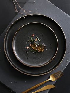 a black plate topped with food on top of a wooden table next to gold utensils