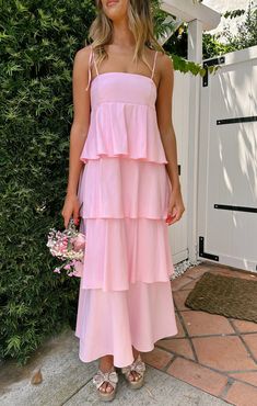 a woman standing in front of a bush wearing a pink tiered dress and sandals