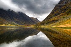 a lake surrounded by mountains with clouds in the sky and water reflecting it's surface