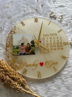 a white clock with gold numbers and a photo on the front is sitting next to a dried plant