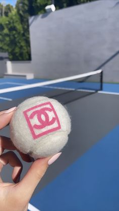 a person holding up a tennis ball with the word chanel painted on it in front of a tennis court