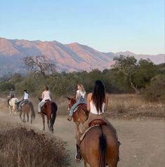 several people riding horses on a dirt road in front of some hills and trees with mountains in the background