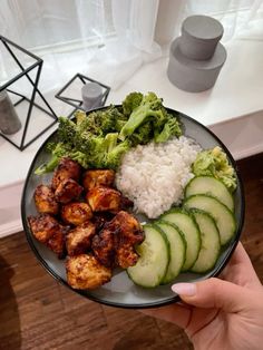 a person holding a plate with chicken, broccoli and rice on it in front of a window