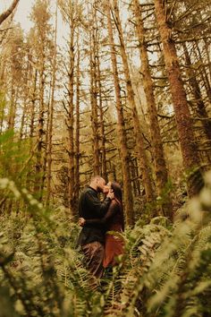 a man and woman standing in the middle of a forest hugging each other with trees behind them