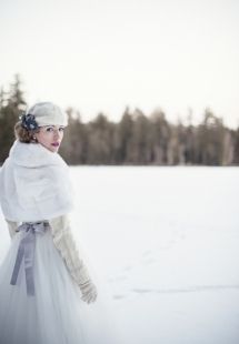 a woman in a white dress and fur coat standing in the snow with trees behind her