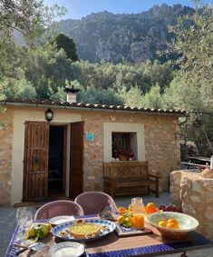 an outdoor table with plates of food on it in front of a stone building and mountains