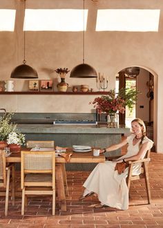 a woman sitting at a wooden table in front of an outdoor kitchen with potted plants