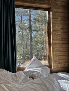 an unmade bed in front of a large window with forest outside the window and blue curtains