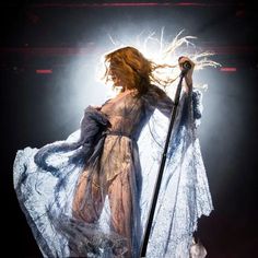 a woman in a white dress holding a microphone and standing on stage with her hair blowing in the wind