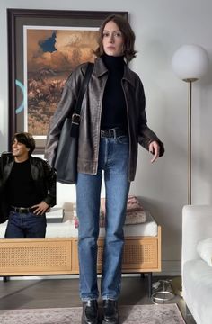 a woman standing in front of a couch wearing jeans and a leather jacket with a black turtle neck sweater