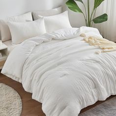 a bed with white comforters and pillows in a room next to a potted plant