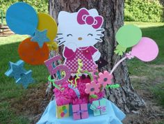 a hello kitty birthday cake on a table with balloons and presents in front of a tree