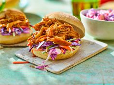 two pulled pork sandwiches with coleslaw slaw on a cutting board next to a bowl of coleslaw