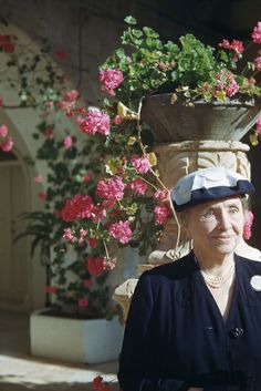 an older woman standing in front of pink flowers with a potted plant behind her