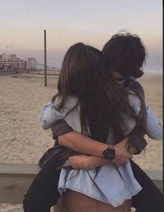 two people hugging each other while sitting on a bench at the beach in front of the ocean