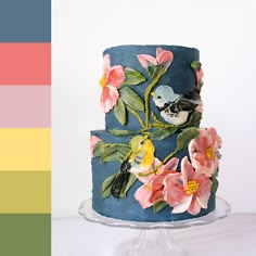 a three tiered cake decorated with flowers and birds on blue icing, sitting on a clear plate