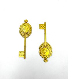 two golden key shaped brooches on a white surface, one has a gold medallion and the other is a diamond