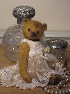 a brown teddy bear wearing a white dress sitting on a table next to beads and a glass jar