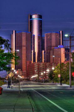 a city street with tall buildings in the background at night, lit up by street lamps