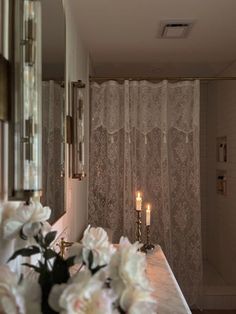 candles are lit on the counter in front of a mirror with flowers and curtains behind it