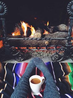 someone sitting in front of a fireplace with their feet up on the floor and holding a cup of coffee