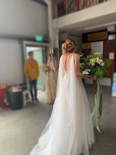 a woman in a wedding dress is standing near a bookshelf and holding flowers